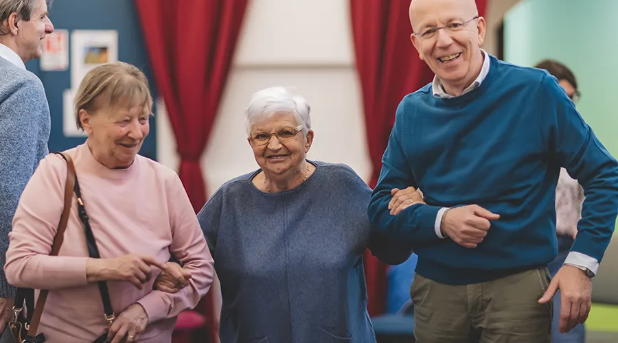 Tre familiari e caregiver nell'Alzheimer café sottobraccio insieme, sorridono.