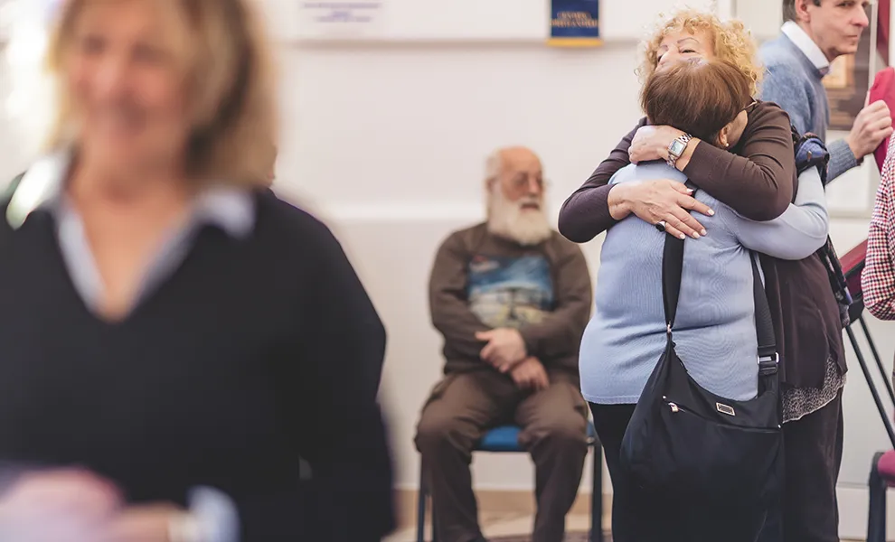 Tra le tante persone presenti nella sede dell'Alzheimer Café, due signore si abbracciano affettuosamente.