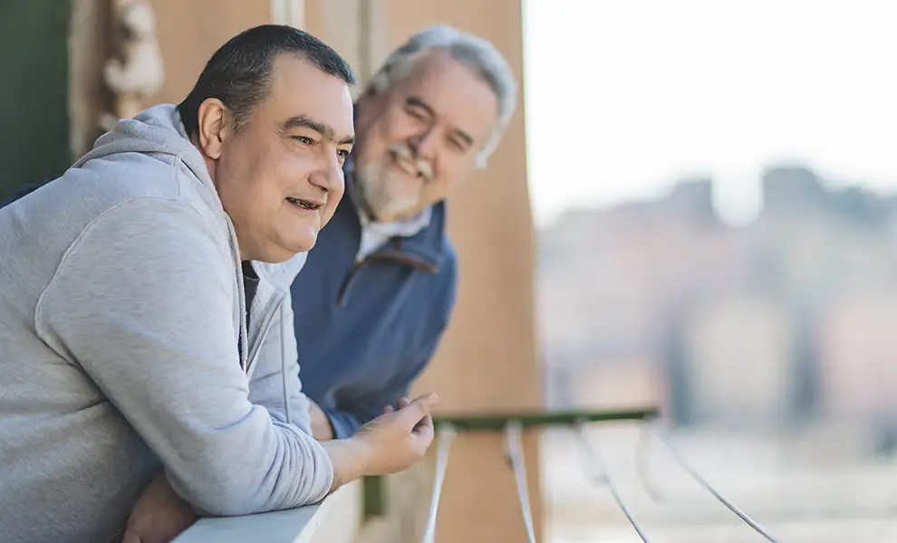 Un ospite ed un medico della fondazione si affacciano sul balcone sorridenti. Davanti a loro la vista dall'alto della città di Genova.